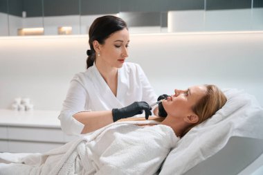 Kozmetik kliniğinde doktor estetisyen kadın çenesine güzellik iğnesi yapar, hasta kanepede yatar.