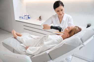 Modern bir kozmetoloji kliniğinin çalışanı güzellik iğnesi yapar, hasta özel bir kanepede yatar.