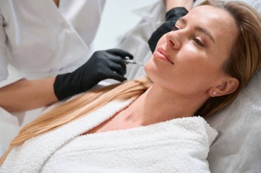 Güzel bir kadın çenesine gençleştirici iğne vuruluyor. Doktor koruyucu eldivenlerle çalışıyor.