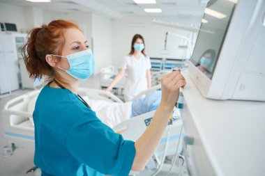 Hayati fonksiyonlarını kontrol eden kadın doktor monitörde, meslektaşı hasta yatağının yanında dikilirken.