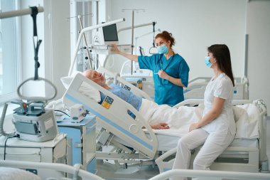 Female doctors in medical mask visiting their male patient who lying in the bed in the hospital ward clipart