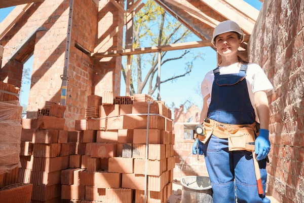 建設現場に沿って歩き バケツでモルタルを運び レンガを敷設するための材料を準備する均一な 硬い帽子と工具ベルトの女性ビルダー — ストック写真