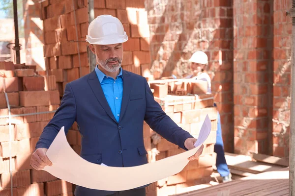 Serious man lead architect in business suit and hardhat looking at building blueprint standing at construction site, checking building quality