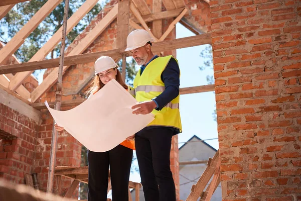 建設中の建物の中に立っている床計画を見て硬帽子の建設労働者 用語や材料を議論 — ストック写真