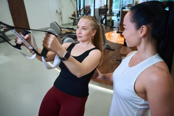 强壮的女人在一个年轻的健身教练旁边做着一个现代的模拟器 — 图库照片