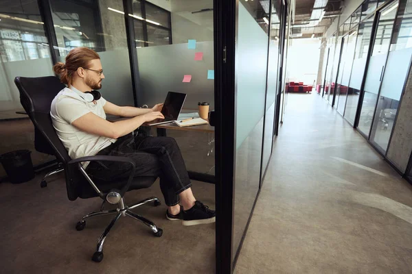 彼の首の周りにヘッドセットを持つ若い従業員は 快適なオフィスブロックに座って 彼はラップトップで動作します — ストック写真