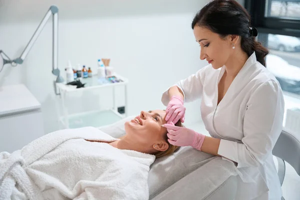 美容師は 患者の顔に作業領域をマークするために特別なマーカーを使用し 女性は美容室ソファに位置しています — ストック写真