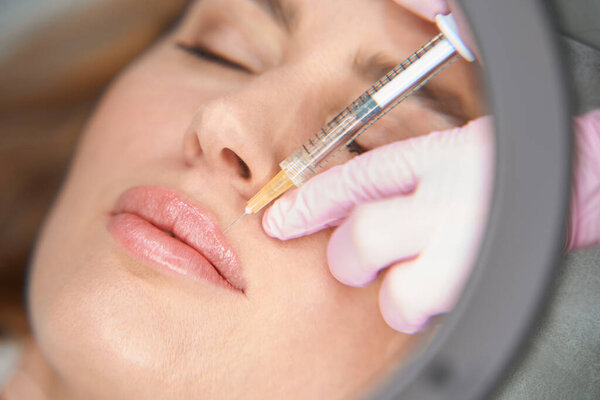 Эстетик делает инъекции женщине в носогубную складку, специалист использует увеличительное стекло