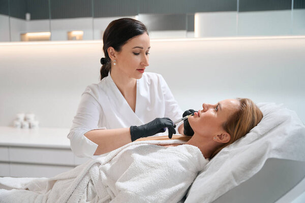 Врач-эстетик в косметологической клинике делает инъекцию красоты в подбородок женщины, пациент лежит на диване