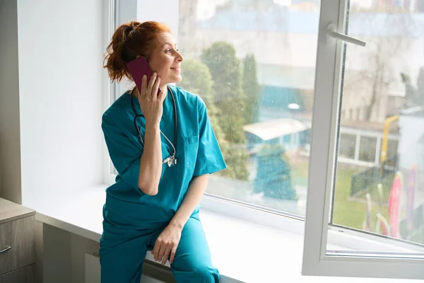 Kvinnlig Läkare Med Stetoskop Att Samtal Smartphone Medan Sitter Fönsterbräda — Stockfoto