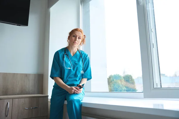 Allvarlig Kvinnlig Läkare Medicinsk Uniform Vilar Fönsterbrädan Efter Hård Arbetsdag — Stockfoto