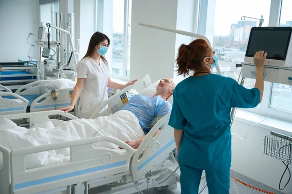 患者のベッドの横に彼女の同僚が立っている間 女性医師は モニター上の必須生理学的指標の規制を制御します — ストック写真