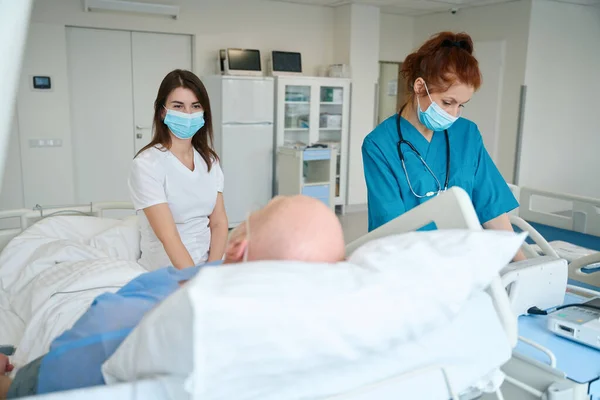 老人と一緒にベッドの近くに立って 病院病棟で彼の健康について学ぶ専門家の女性の専門家 — ストック写真