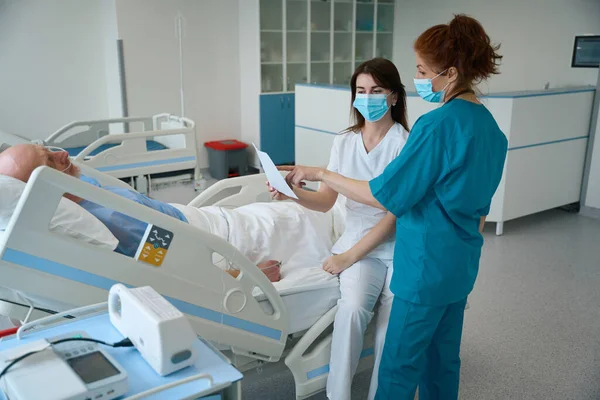 病院のベッドに寝そべっている男性患者と話す紙のシートを持つ制服の女性医師 — ストック写真