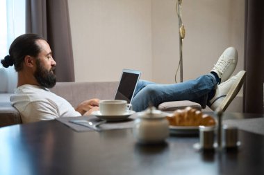 Orta yaşlı erkek, yumuşak bir kanepede dizüstü bilgisayarla oturuyor. Yakın bir masadaki masaya hafif bir kahvaltı konuyor.