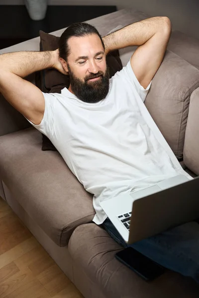 よく整形されたひげを持つ男は ラップトップ付きのソファーで休んでいる 彼は快適にレクリエーションエリアに位置しています — ストック写真