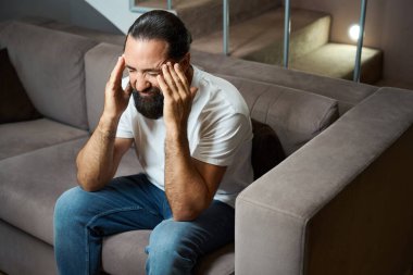 Sakallı adam baş ağrısı çekiyor, yumuşak bir kanepede oturuyor.