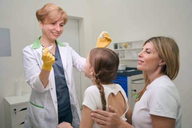 Bir kız göz doktoruyla randevusunda annesinin kucağına oturur. Doktorun elinde ilaç olan bir pipet vardır.