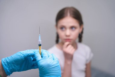 Koruyucu eldivenli doktor elinde ilaç olan bir şırınga tutuyor. Genç bir hasta tıbbi kanepede oturuyor.