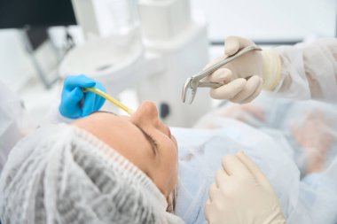Diş çekmek için dişçi koltuğunda oturan bir kadın özel pens ve tükürük püskürtücü kullanır.