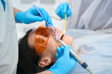 Doktor, hastaların dişleriyle ilgilenen bir asistanın yardımıyla, kadın gözlük takıyor.