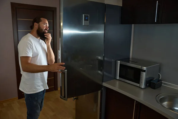 ジーンズのひげ付き男は大きな冷蔵庫から何かを取り出し その隣には電子レンジがあります — ストック写真