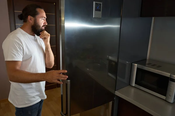 中年男性は 電子レンジの隣にある冷蔵庫の前に立って考えます — ストック写真
