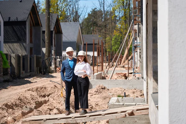 Positive woman general designer and foreman in hardhats smiling standing near builded house, satisfied with construction compliance, going to finish in time