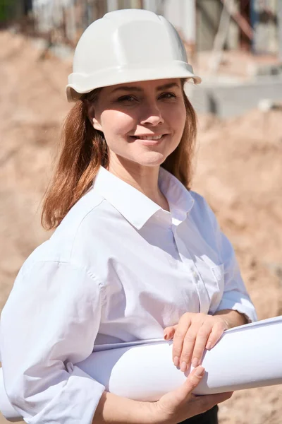 Portrait of beautiful woman architect or landscape designer in hardhat standing at construction site holding building project at hands, success