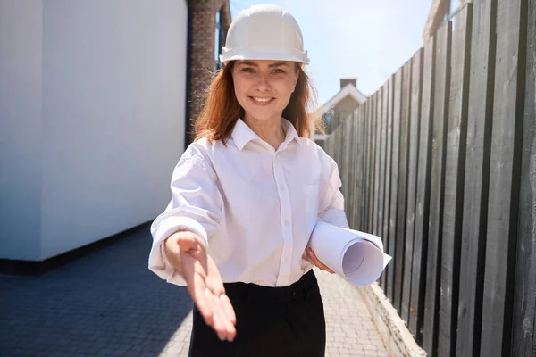 穿着硬礼帽的女建筑师微笑欢迎承包商 伸出手来握手 拿着建筑蓝图讨论细节 — 图库照片