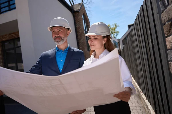 Woman architect showing blueprint to general contractor, asking for advice and discussing detail before building start, coming at construction site