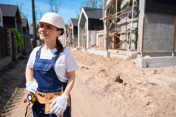 建設現場に立っている堅くて工具ベルトの自信ある女性総合請負業者 建築資材の作業範囲と品質を評価 — ストック写真
