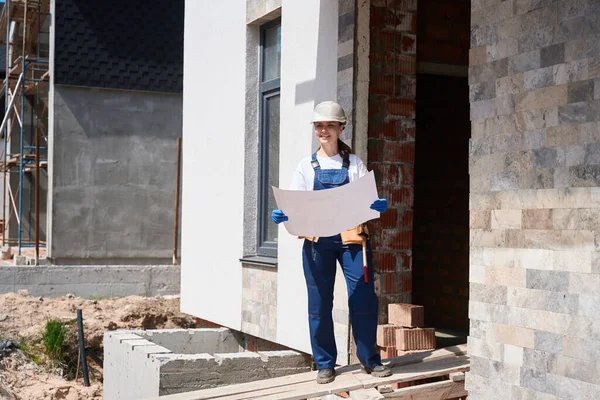建設エントランス下の家の近くに立つポジティブな女性請負業者で ブループリントの建設 仕上げ作業の計画 — ストック写真