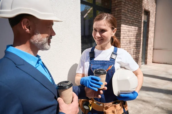 Man general contractor treating workwoman in overall with coffee, colleagues sweetly talking at construction site during time-break, friendly relations, flirt