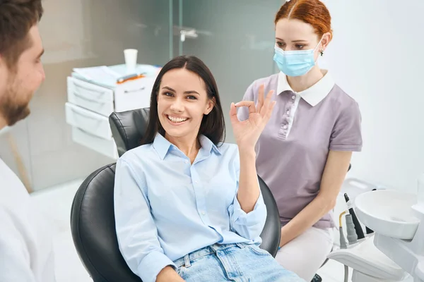 歯科検査およびクリーニングの後で正統な椅子に座ってOkの印を示す専門の歯の処置の女性の顧客に満足して下さい — ストック写真