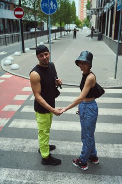 Neşeli adam ve kadın kameraya bakıyor ve yaya geçidinde dururken el ele tutuşuyorlar.