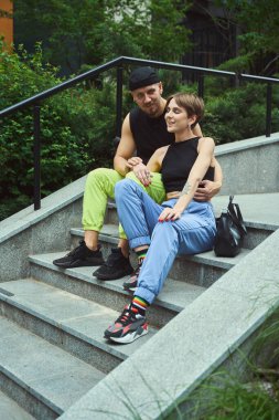 Genç adam merdivenlerde otururken kameraya bakıyor. Parktaki kız arkadaşının yanında.