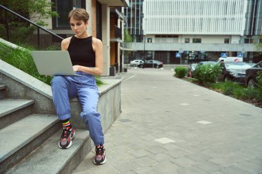 Modern caddede çimlerin yanında oturan şık giysili güzel bir kadın dizüstü bilgisayar kullanıyor.