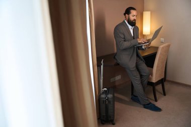 Seyahat eden iş adamı, otel odasının çalışma alanında dizüstü bilgisayarla iletişim kuruyor. Bir seyahat çantasının yanında.