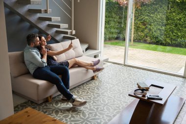 Orta yaşlı bir çift, geniş bir salonda kucaklaşarak oturur ve selfie çekerler.