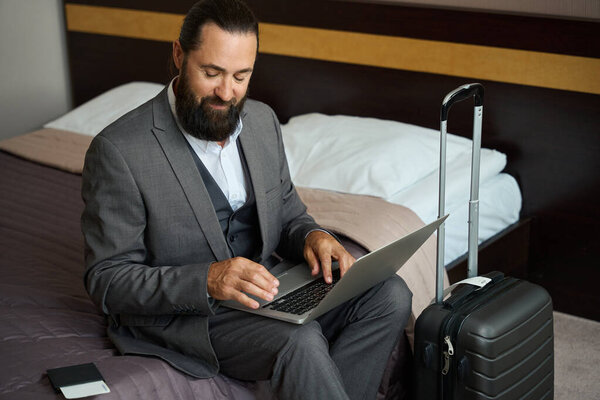 Traveling man sat down with a laptop on the bed, next to his passport and ticket
