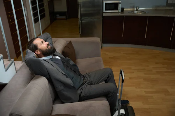 道からの男はホテルの部屋の柔らかいソファーで休んでいる 彼は旅行のスーツケースを持っています — ストック写真
