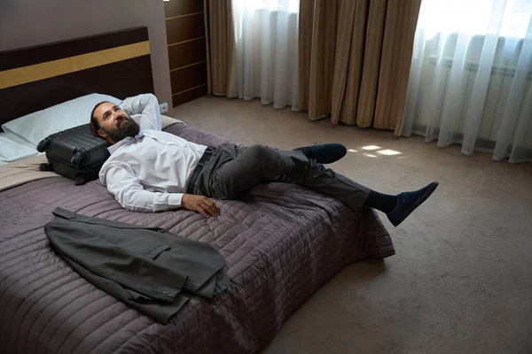 ひげ付きの男は大きなベッドの上の道路から休むために横たわり 部屋はきれいで快適です — ストック写真