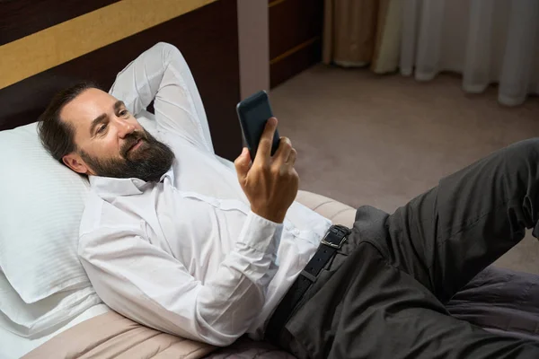 Tired male reads the news in a mobile phone, he lies on a big bed