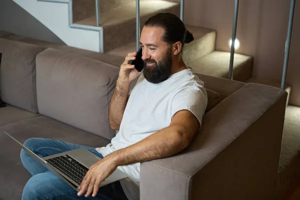 Smiling man communicates on a mobile phone and works on a modern laptop, he is in comfortable casual clothes