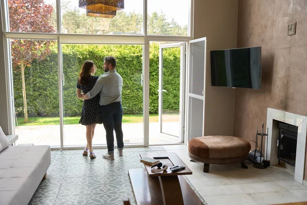 Middle-aged couple is dancing, embracing, in a spacious living room, outside the window is a cute backyard