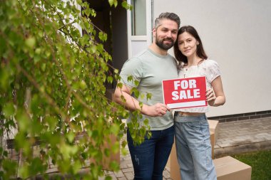 Genç bir adam ve kadın bahar bahçesinde dikilirken kameraya bakıyor ve satılık tabela tutuyor.