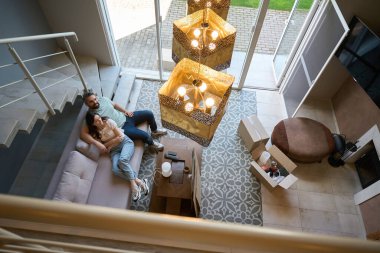 Yatakta uzanan bir kadın ve yanında oturan ve kapalı alanda kameraya bakan bir adam.