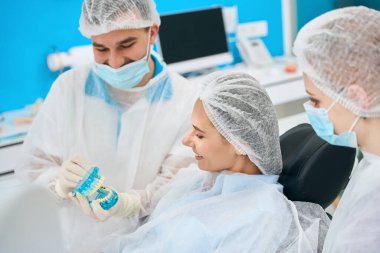 Kadın, diş implantlarıyla bir maketi inceliyor, doktor ve asistan koruyucu maskeler kullanıyor.