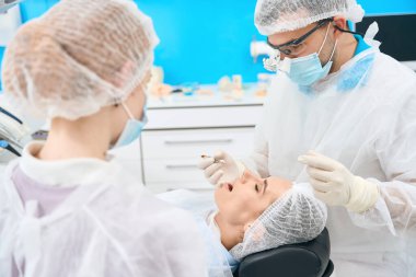 Diş hekimi, diş kanalını mikroskop altında genç bir kadına, tıbbi üniformalı bir asistana mühürlüyor.
